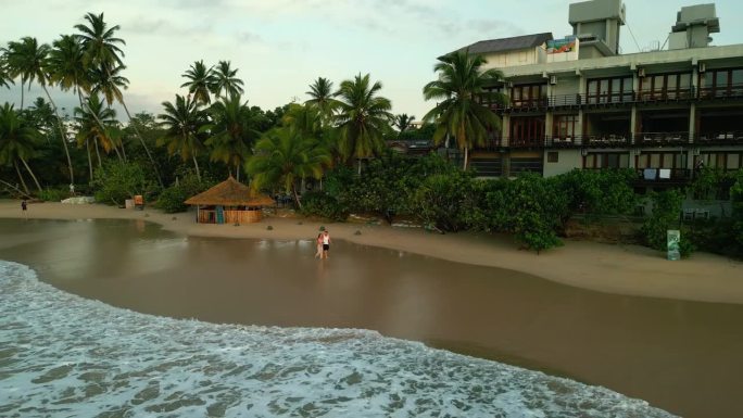 一对夫妇手牵着手走在热带海滩上，在日出的海洋边看无人机镜头。男友和女友在海边沙滩上靠近水，拥抱，亲吻