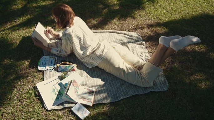 一个充满灵感的女人在阳光明媚的日子里躺在公园的毯子上，在速写本上画画