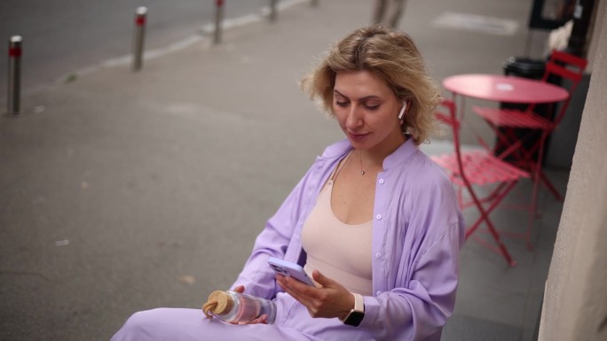 一头卷发的年轻金发女郎坐在街边的咖啡馆里，一边听着音乐，一边用智能手机浏览手机应用