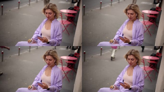 一头卷发的年轻金发女郎坐在街边的咖啡馆里，一边听着音乐，一边用智能手机浏览手机应用
