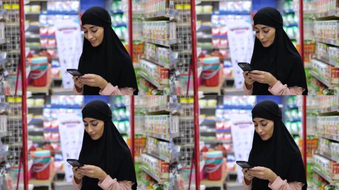 一名戴着头巾的美女站在商店的货架中间，手里拿着手机，微笑着回答留言。女人喜欢和她的对话者在电话中交谈