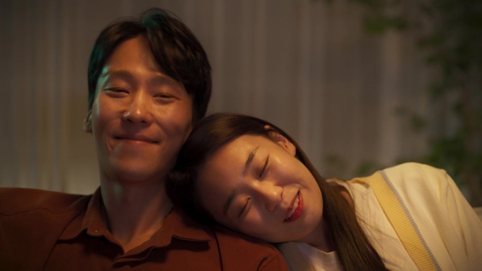 年轻的亚洲夫妇坐在家里的沙发上通过在线流媒体服务观看电影。韩国男人和美丽的女友一起享受浪漫的电视节目