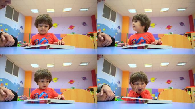 幼儿园室内，一个男孩坐在桌子旁看故事书，看着镜头