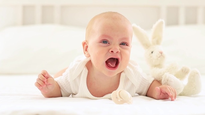 在明亮的卧室里，一个穿着白色紧身衣的婴儿在床上哭泣，一个蓝眼睛的可爱婴儿趴在地上叫他的妈妈，一个孩子