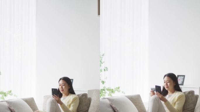 竖屏:手持信用卡使用智能手机的韩国女性。亚洲女性做移动购物疗法，在客厅浏览网上购物网站，发现时尚趋势
