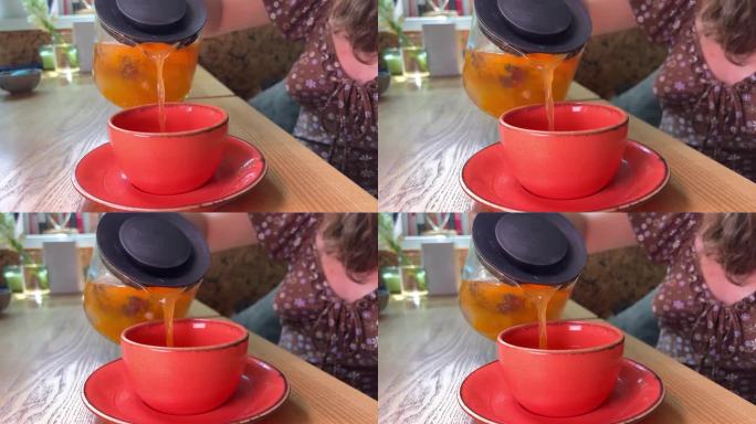沙棘茶从一个玻璃茶壶倒进一个亮粉色的杯子在一个盘子木桌子上的女孩在一个棕色的裙子认不出来的人美味的饮