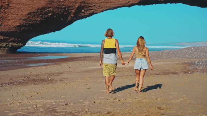 一对幸福的夫妇在沙滩上散步，手牵着手走向红砂岩天然拱门