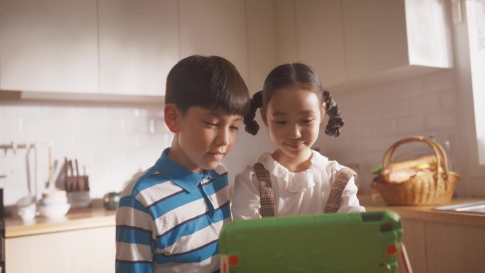 两个韩国孩子周末在厨房里使用电子平板电脑的照片。两个可爱的亚洲兄弟姐妹在线观看教育内容，分享小工具，