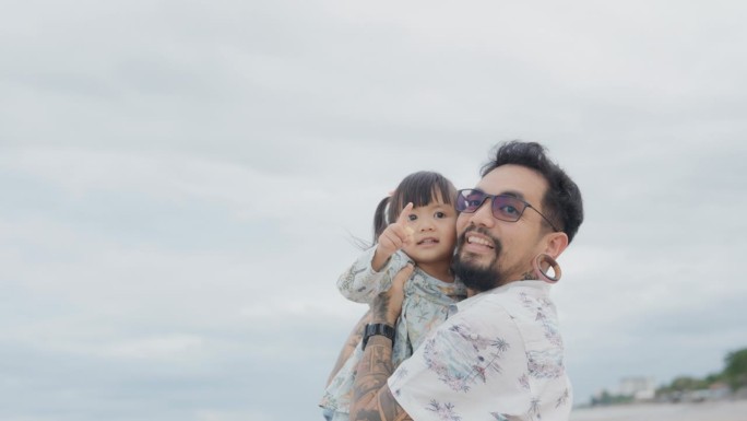快乐的亚洲父亲，抱着小女儿，看着大海，深情地亲吻她的脸颊。