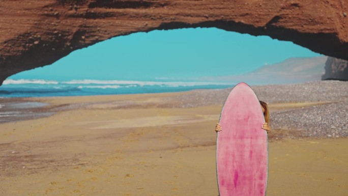 年轻女子站在红色砂岩地层前的海滩上，站在粉色冲浪板后面望向镜头