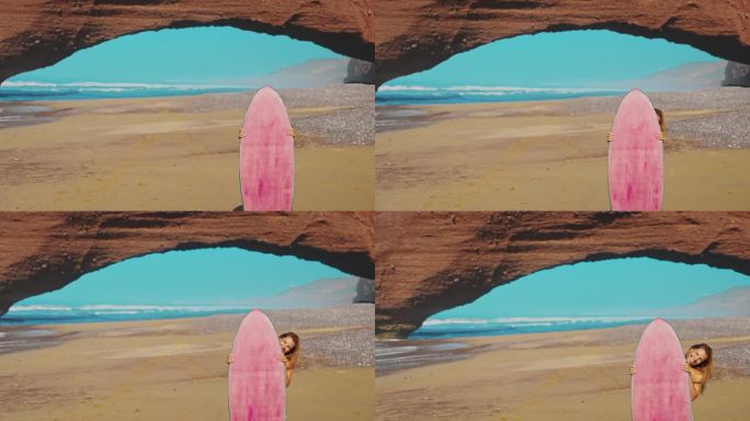 年轻女子站在红色砂岩地层前的海滩上，站在粉色冲浪板后面望向镜头