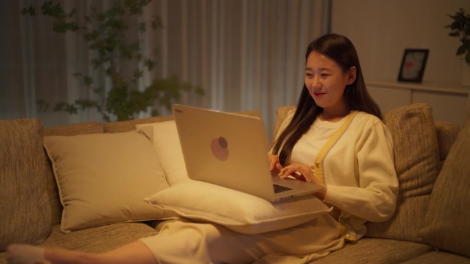 韩国女性深夜在家专注于虚拟工作，舒适地坐在沙发上，使用笔记本电脑进行创意写作和为国际团队制作演示文稿