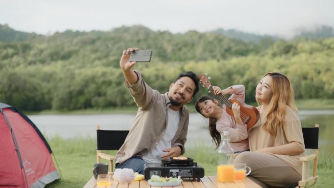 幸福的一家人在绿地上露营，父亲坐在野餐桌旁用智能手机自拍。