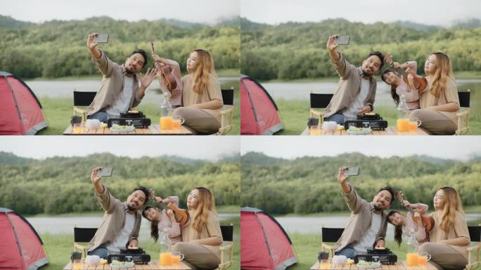 幸福的一家人在绿地上露营，父亲坐在野餐桌旁用智能手机自拍。