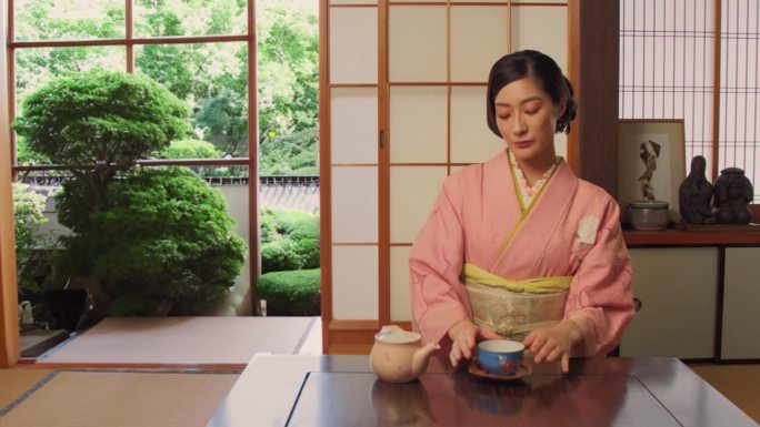 年轻的日本女性享受独自在家喝茶的时光。漂亮的亚洲女人穿着经典的粉红色和服，坐在一个传统的日本家庭的矮