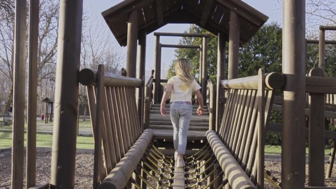 阳光明媚的一天，一个白人小女孩在操场上的桥上玩耍。童年与家人在户外玩耍。