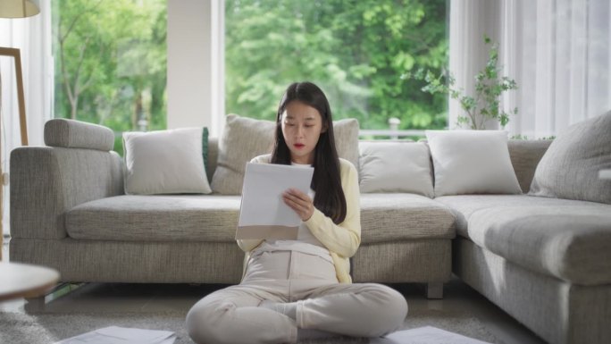 一位韩国妇女坐在舒适沙发旁边的地板上，做大学作业，阅读笔记本上的信息。年轻女性在一个时髦的客厅里写她