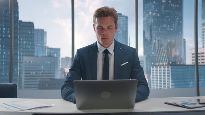 西装革履的自信商人坐在办公室的办公桌前，使用笔记本电脑，靠窗眺望摩天大楼的大城市。成功的财务经理计划