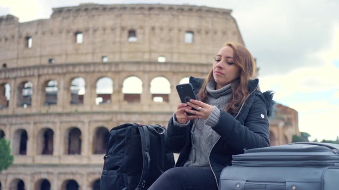 在意大利罗马的历史建筑斗兽场前，带着行李和保暖衣服的年轻拉丁女游客在多云的天空中玩着智能手机