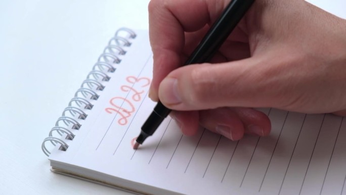 手特写，用粉红色的毡尖笔在笔记本上写下2023年的数字和来年的目标。