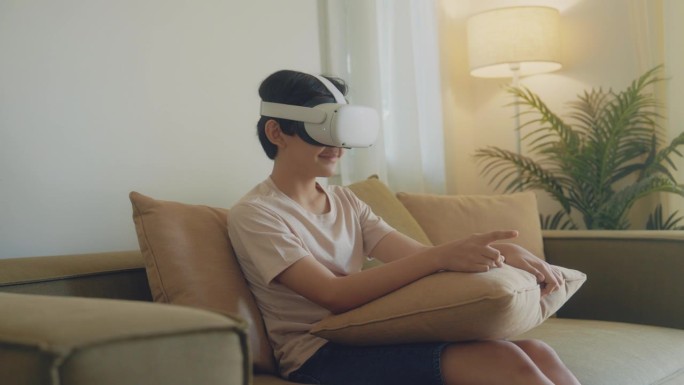 亚洲男孩坐在沙发上，用虚拟现实未来科技耳机在家里玩模拟3D视频游戏。