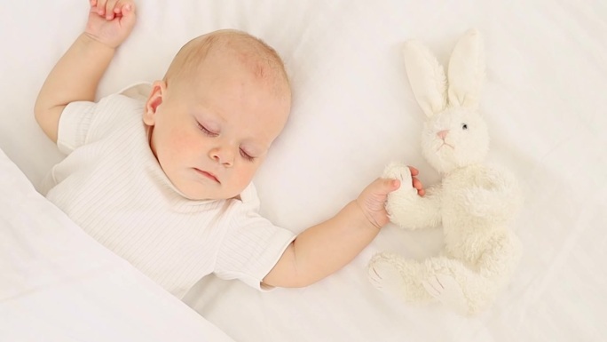 可爱的小新生宝宝抱着毛绒兔子睡在毯子下，甜蜜健康的宝宝睡在白色的婴儿床上，一个文字的地方