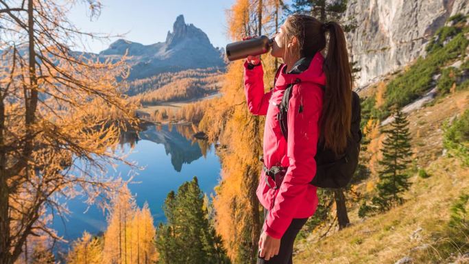 女人在山腰喝水，同时俯瞰雄伟的山间湖泊在秋天