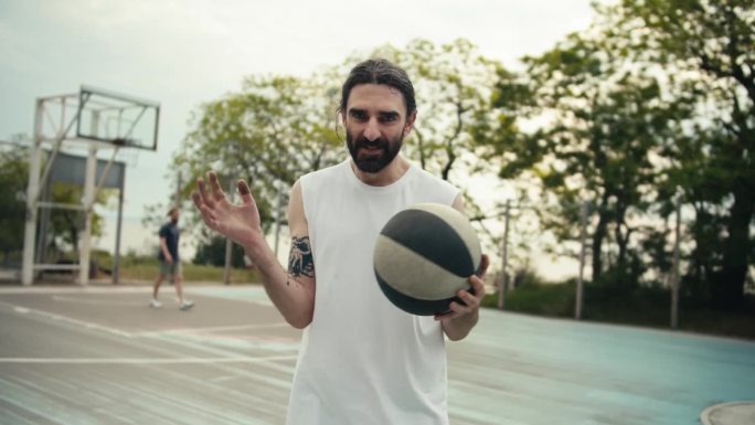 一名身穿白色t恤、留着胡子的中年男子手持篮球，在篮球场的背景下摆姿势