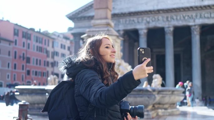 在意大利罗马的万神殿前，穿着保暖衣服的年轻拉丁女子站在智能手机上用专业相机自拍，目光移开