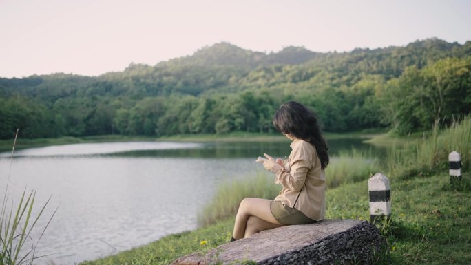 年轻的亚洲女子在大自然的河岸用智能手机拍摄令人惊叹的景色。她在欣赏美丽的景色。美丽的女孩深呼吸，