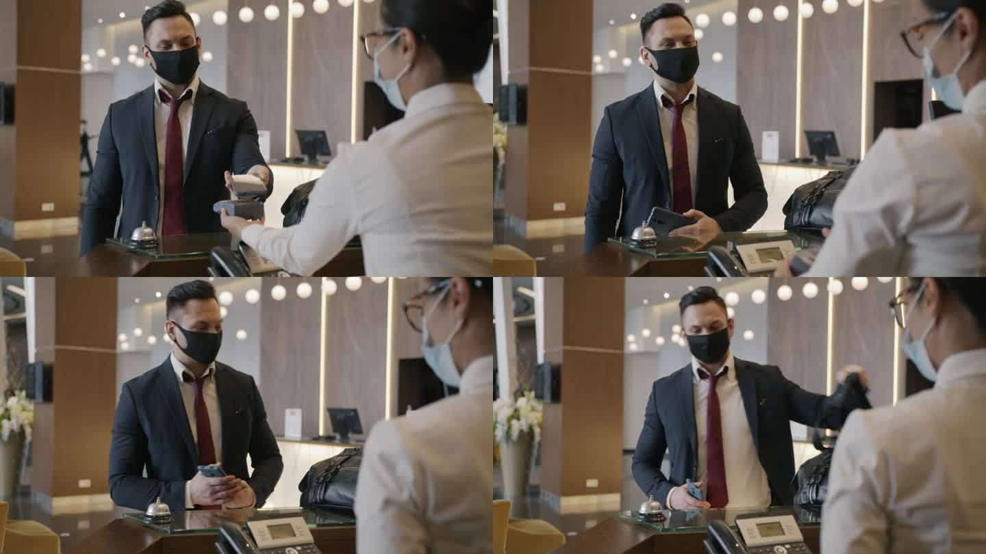 戴着口罩的年轻企业家用智能手机支付酒店服务费用，并与接待员交谈