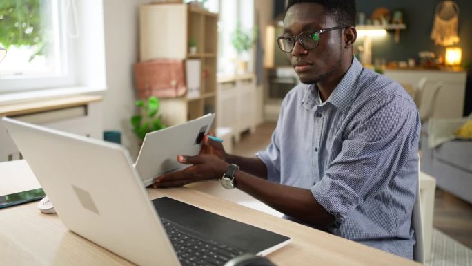在大学宿舍里，一名非裔美国男学生一边用笔记本电脑上网学习