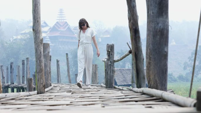 在雾蒙蒙的早晨，一名身穿白衣的年轻女子走在一座木桥上，逐渐向镜头靠近