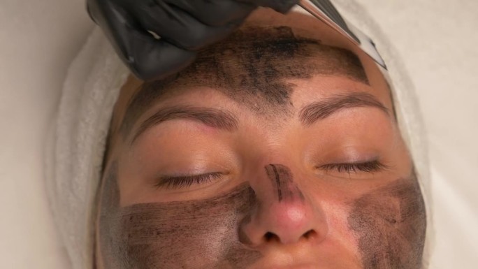 美容师在戴着黑色手套的漂亮女人脸上涂上黑色面膜，漂亮的女人在水疗中心做面部手术