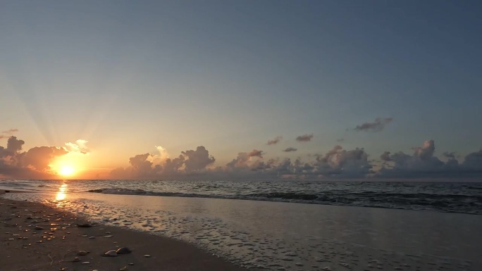 地面水平拍摄的海滩上，太阳升起在海面上，地平线上有云，小波浪拍打着沙滩