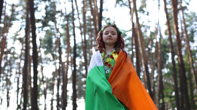 红头发的小女孩身上披着爱尔兰国旗，正穿过森林。可爱的白人小孩在庆祝圣帕特里克节