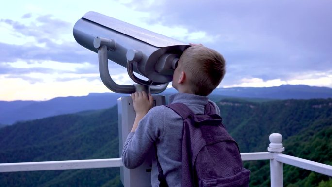 男孩好奇地透过望远镜往外看。山看。用双筒望远镜看天空。和孩子一起旅行。和学生们有趣的旅行。在山上远足