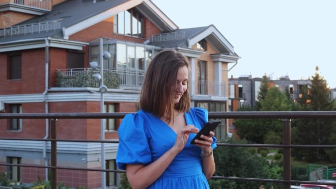 穿着蓝色淡色衣服的年轻女子在城市风景的背景下用智能手机打字