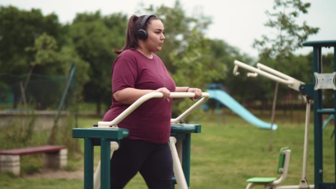 一名戴着耳机的超重年轻女子在室外健身房里的椭圆机上锻炼。