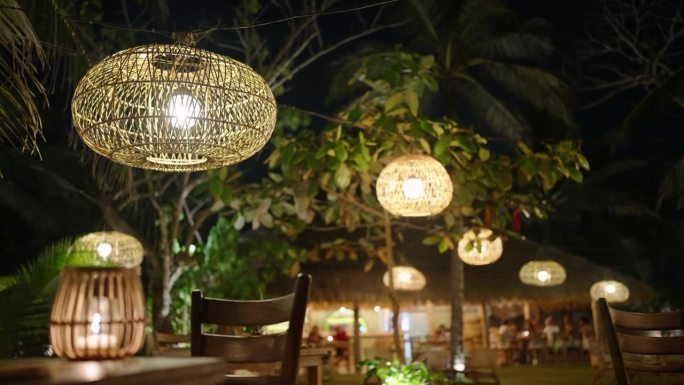 在夜晚的热带背景下，柳条草灯挂在树上作为装饰。老式的木制灯泡在风中摇曳，照亮了户外咖啡馆。波西米亚婚