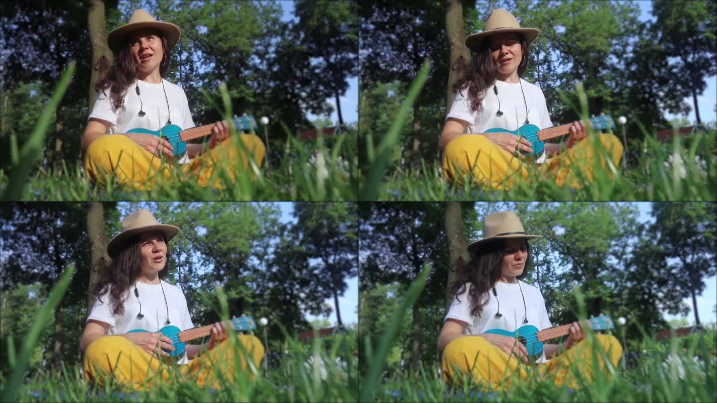 公园里的女孩坐在草地上弹尤克里里。学习演奏四弦乐器。在公园里放松。音乐疗法，心理学，冥想，放松，放松
