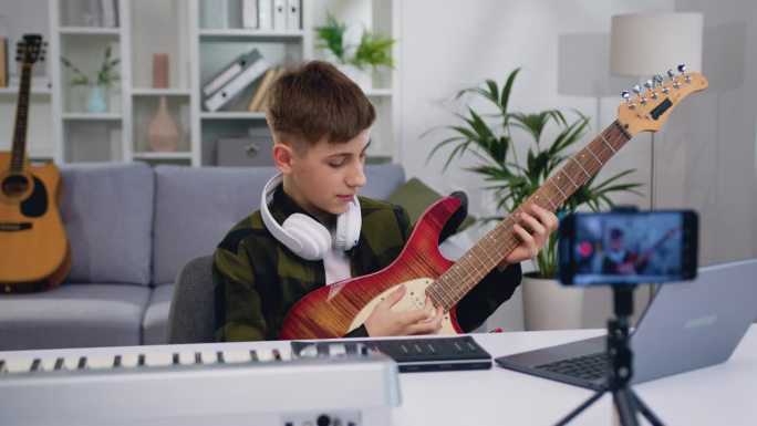 帅气的青少年博主在家庭工作室录制视频课教程，在现代智能手机上弹吉他。十几岁的男孩在网上演示如何弹电吉