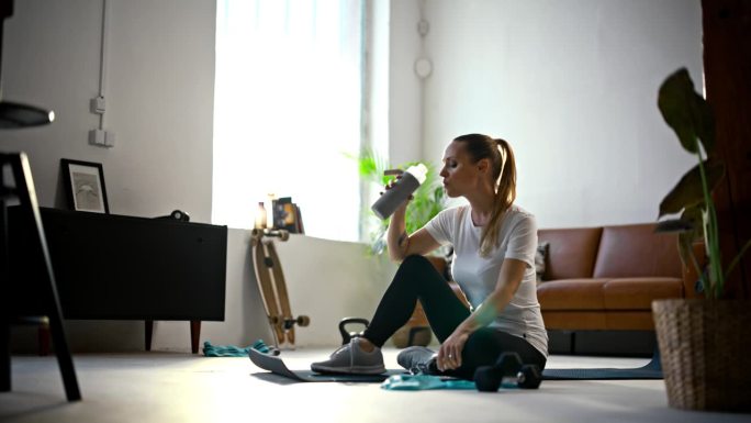 专注的，健康的年轻女子在客厅地板上从水瓶里喝水，在家庭锻炼后休息。运动，健身，健康的生活方式，积极的