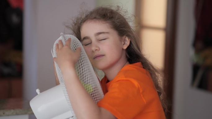 孩子在家里享受电风扇吹来的凉风。女孩在通风机前遭受高温折磨，用电风扇冷却器给自己降温。炎热气候变化。