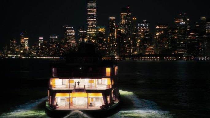夜间，一艘旅游渡轮驶向纽约市摩天大楼的空中无人机镜头。大城市灯光全景城市景观与灯光。深夜曼哈顿下城鸟