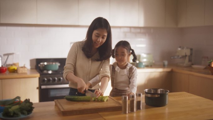 美丽年轻的韩国母亲教她可爱的小女儿如何做饭的慢动作。母亲和女孩子花时间在一起，学习如何安全使用菜刀