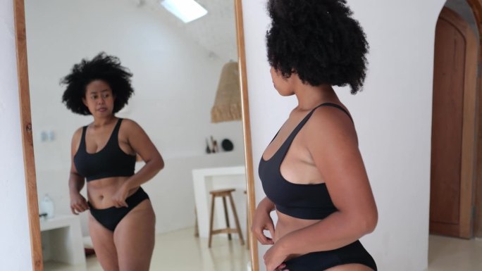 女人看着镜子里的自己，试图接受自己的身体:有妊娠纹，有脂肪团。社会对肥胖身体持有毒批判态度，需要规范