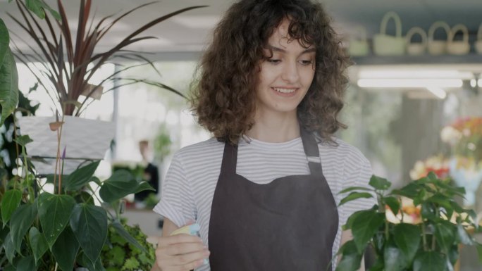 穿着围裙的开朗的年轻女花店老板正在给绿色植物浇水，照顾店里的花