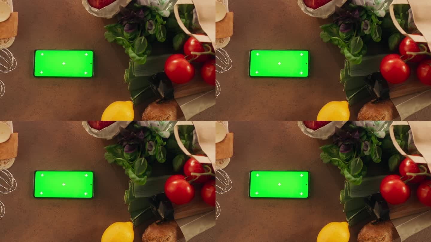 智能手机的顶视图与模拟绿色屏幕显示。一个设备水平躺在厨房桌子上的蔬菜的静态镜头。在线数字营销和内容创