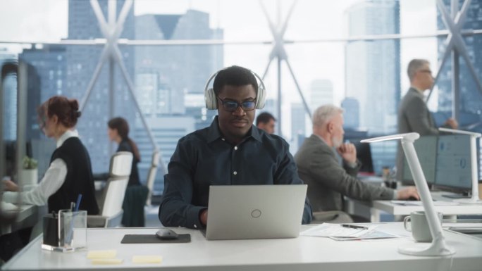 在市中心摩天大楼的办公室里，一个成功的黑人戴上耳机，开始在笔记本电脑上打字。专业的男性产品经理在多样